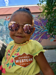 a little girl smiles wearing sunglasses and a National Western Center sticker